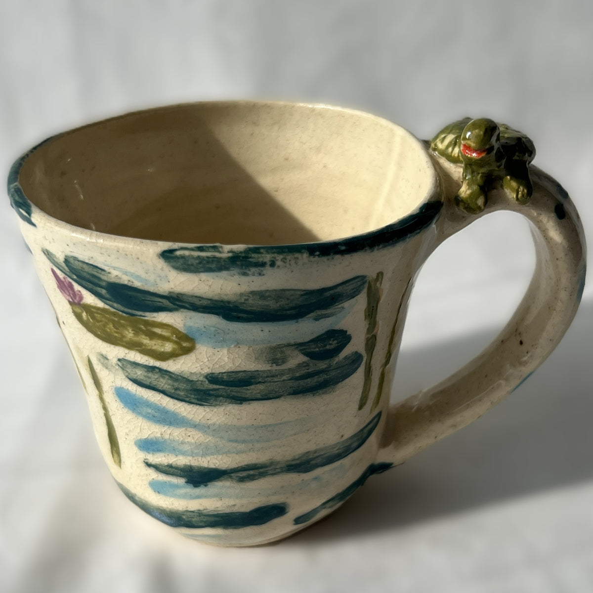 Ceramic mug with a handle featuring artistic patterns on a plain background. There is a small frog statue on the handle
