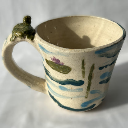Ceramic mug with a handle featuring artistic patterns on a plain background. There is a small frog statue on the handle