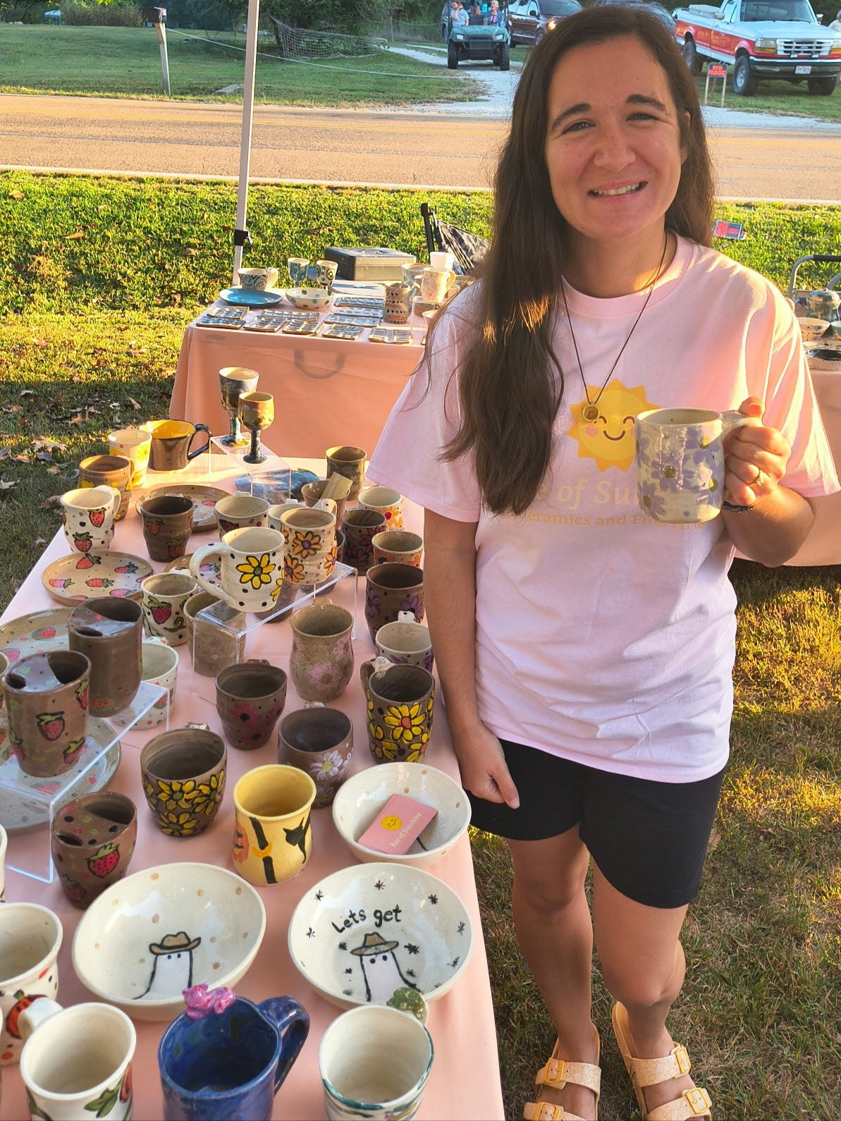 Woman holding a ceramic mug at an outdoor craft fair with tables of handmade items.
